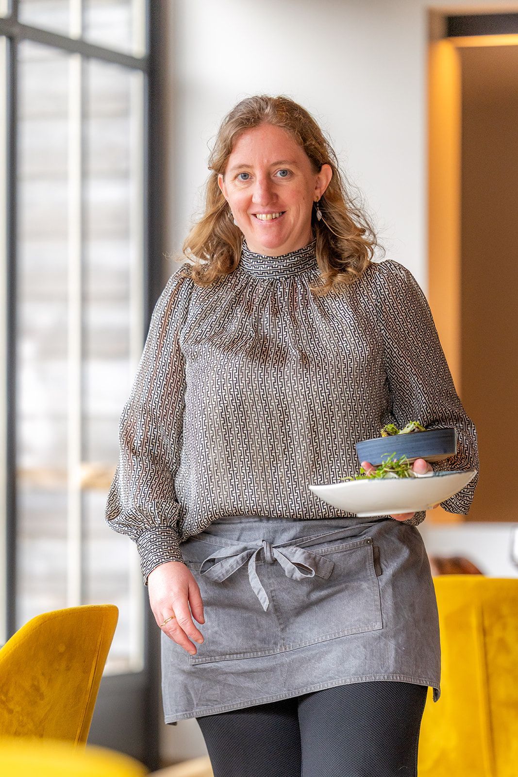 A smiling person stands in a restaurant wearing a patterned blouse and apron, holding two small plates of food.