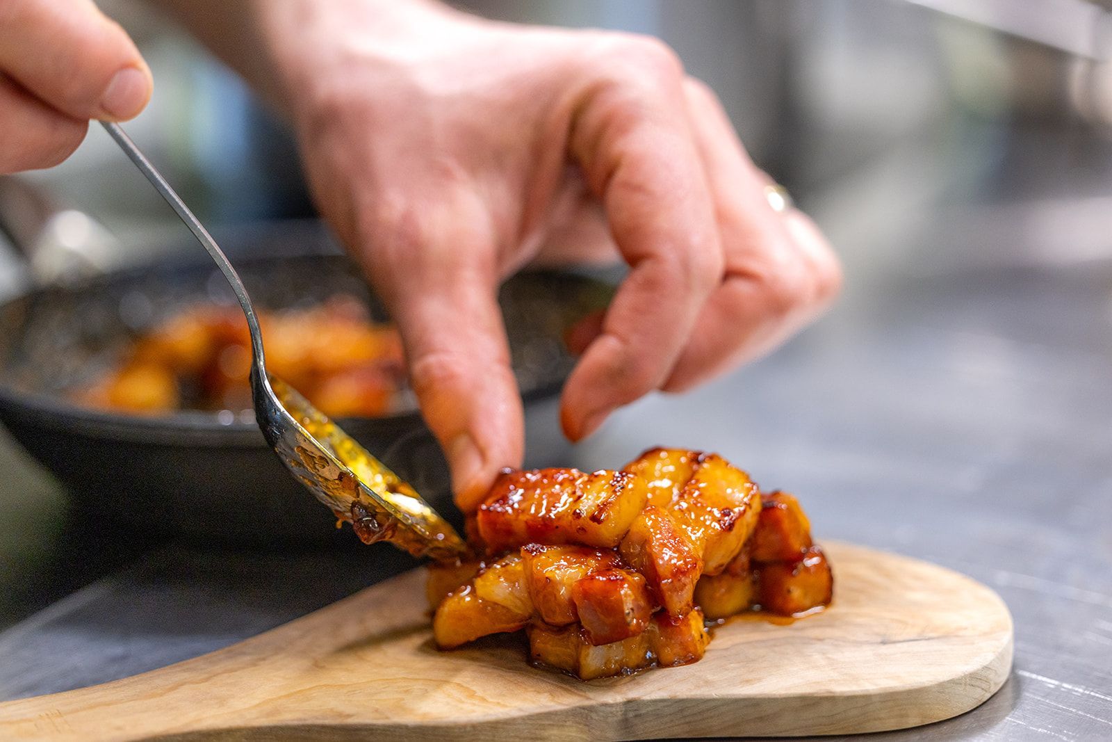A close-up view of a chef’s hands using a metal spoon to arrange glazed, diced vegetables onto a wooden serving board.