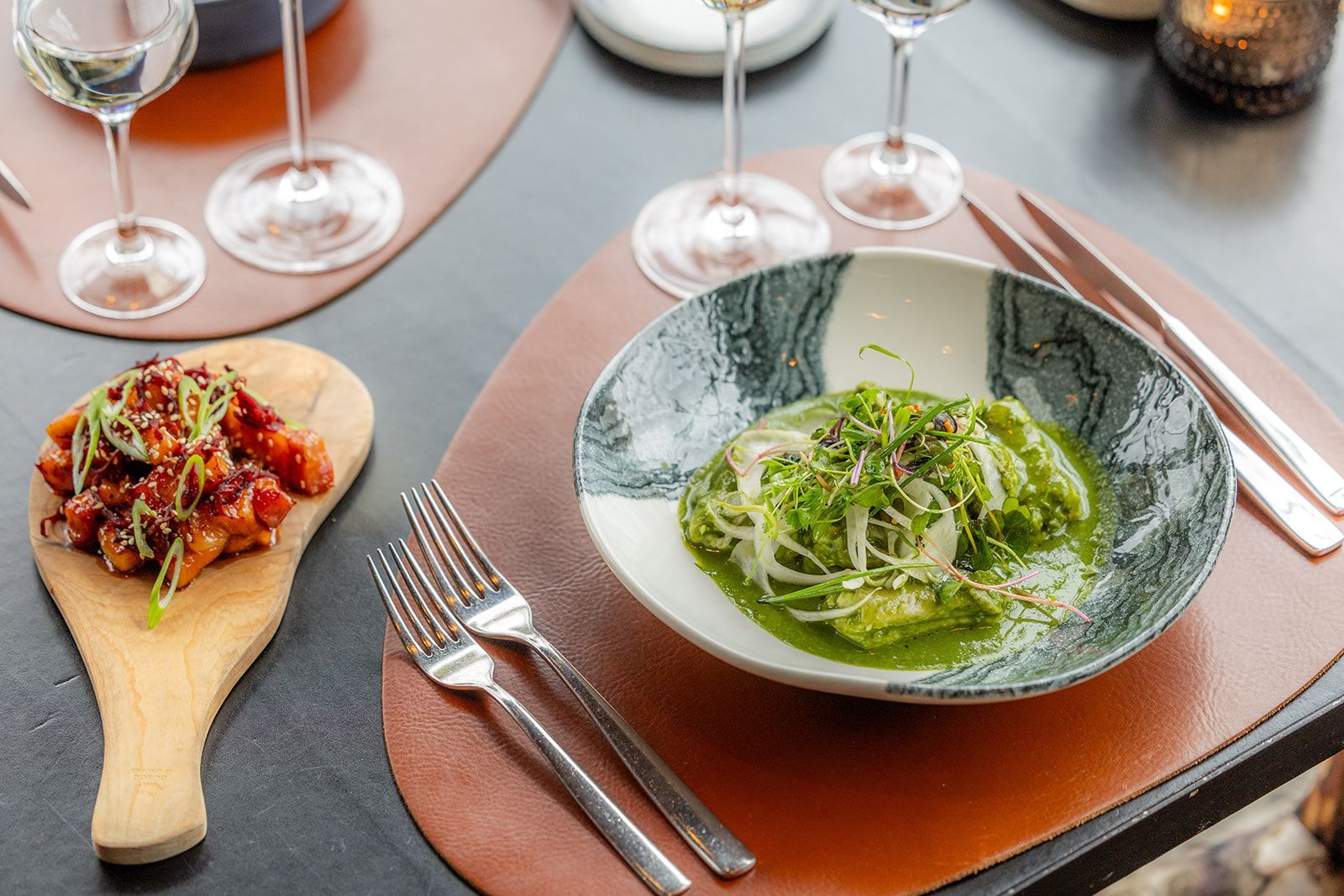 A bowl of green pesto pasta next to a wooden paddle with small appetizers on a leather placemat with glasses.