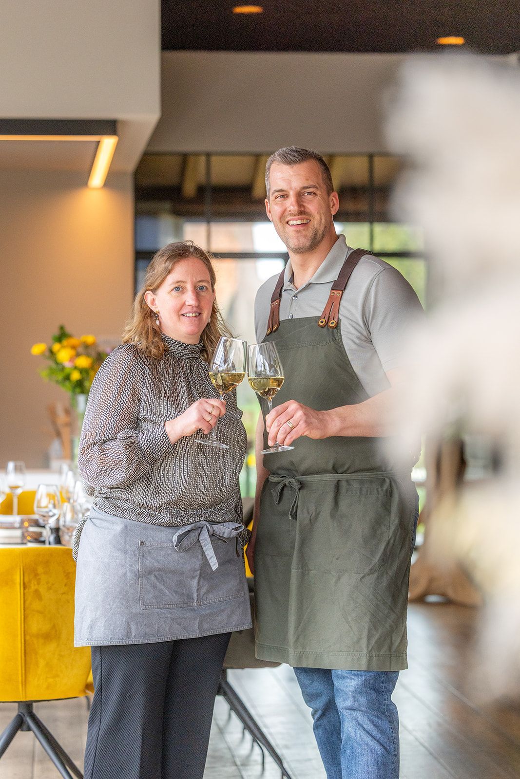 Two staff members in aprons smile and toast with glasses of white wine inside a restaurant.