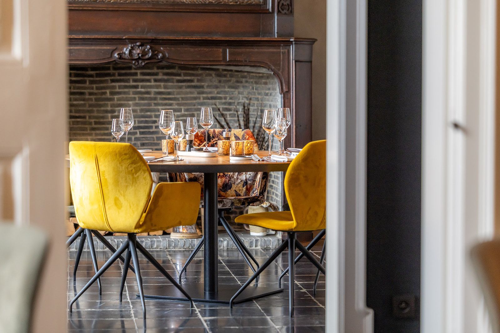 A dining area viewed through a doorway, featuring a round table, two bright yellow velvet chairs, and a stone fireplace.