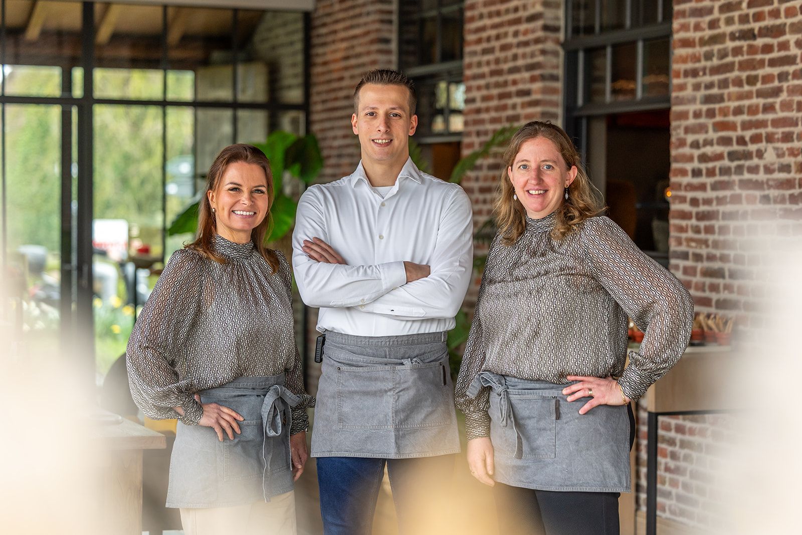 Three smiling staff members in patterned blouses or a shirt and grey aprons stand together inside a brick-walled space.