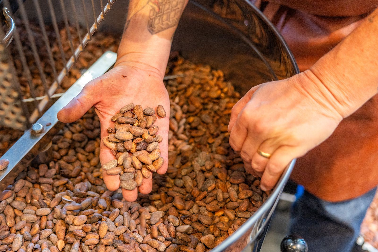 A chef in a dark apron and cap smiles while plating a green sauce from a small pan in a professional kitchen.