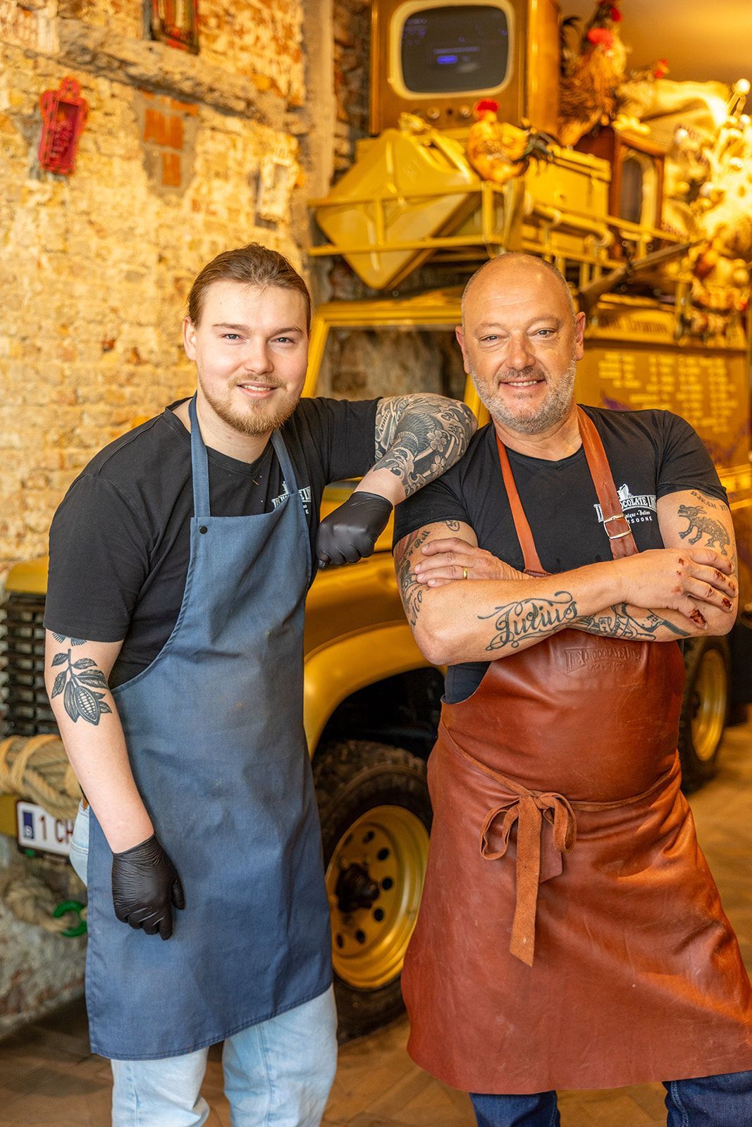 A smiling person stands in a restaurant wearing a patterned blouse and apron, holding two small plates of food.