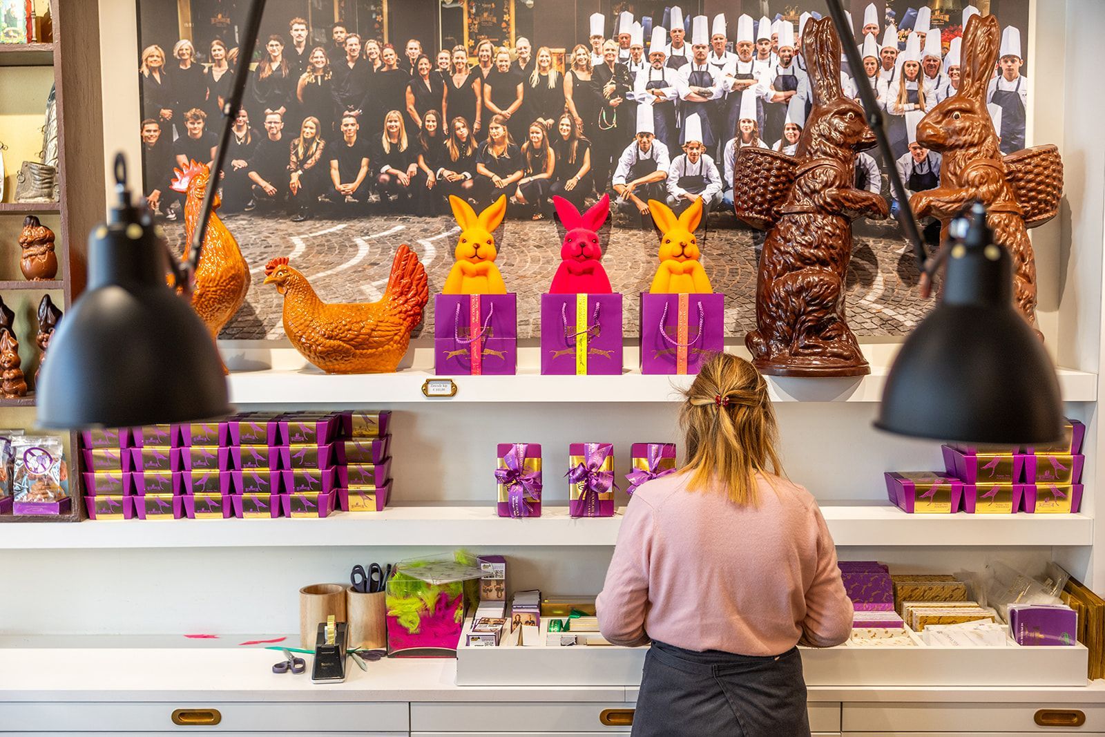 A close-up view of a chefβs hands using a metal spoon to arrange glazed, diced vegetables onto a wooden serving board.