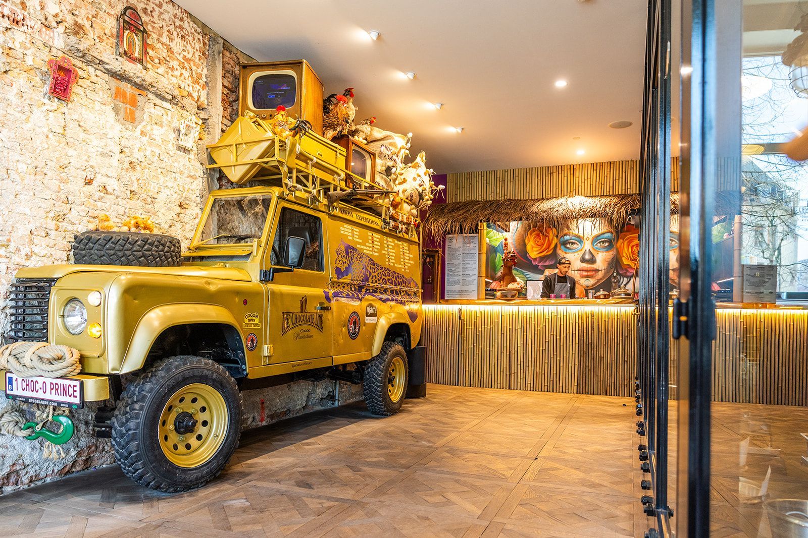 A restaurant interior with two tables, yellow and patterned chairs, brick flooring, and deer head wall decor.