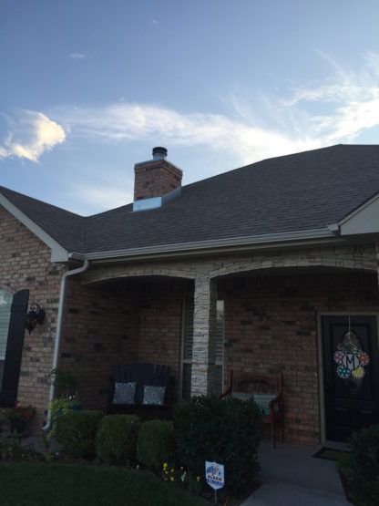 The front exterior of a single-story brick home with a shingled roof, chimney, covered porch, and landscaping.