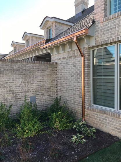 A light-colored brick house featuring a shiny copper gutter and downspout, with dormer windows and small garden plants.