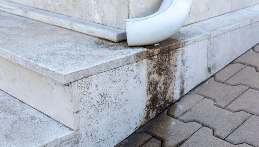 A white downspout discharging water onto light-colored stone steps, creating a dark, vertical stain on the stone surface.