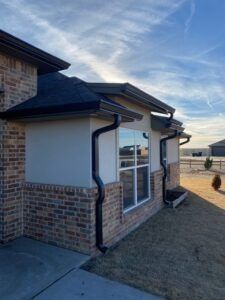 Side view of a house exterior featuring brick and light-colored siding, black roof trim, and two black downspouts.