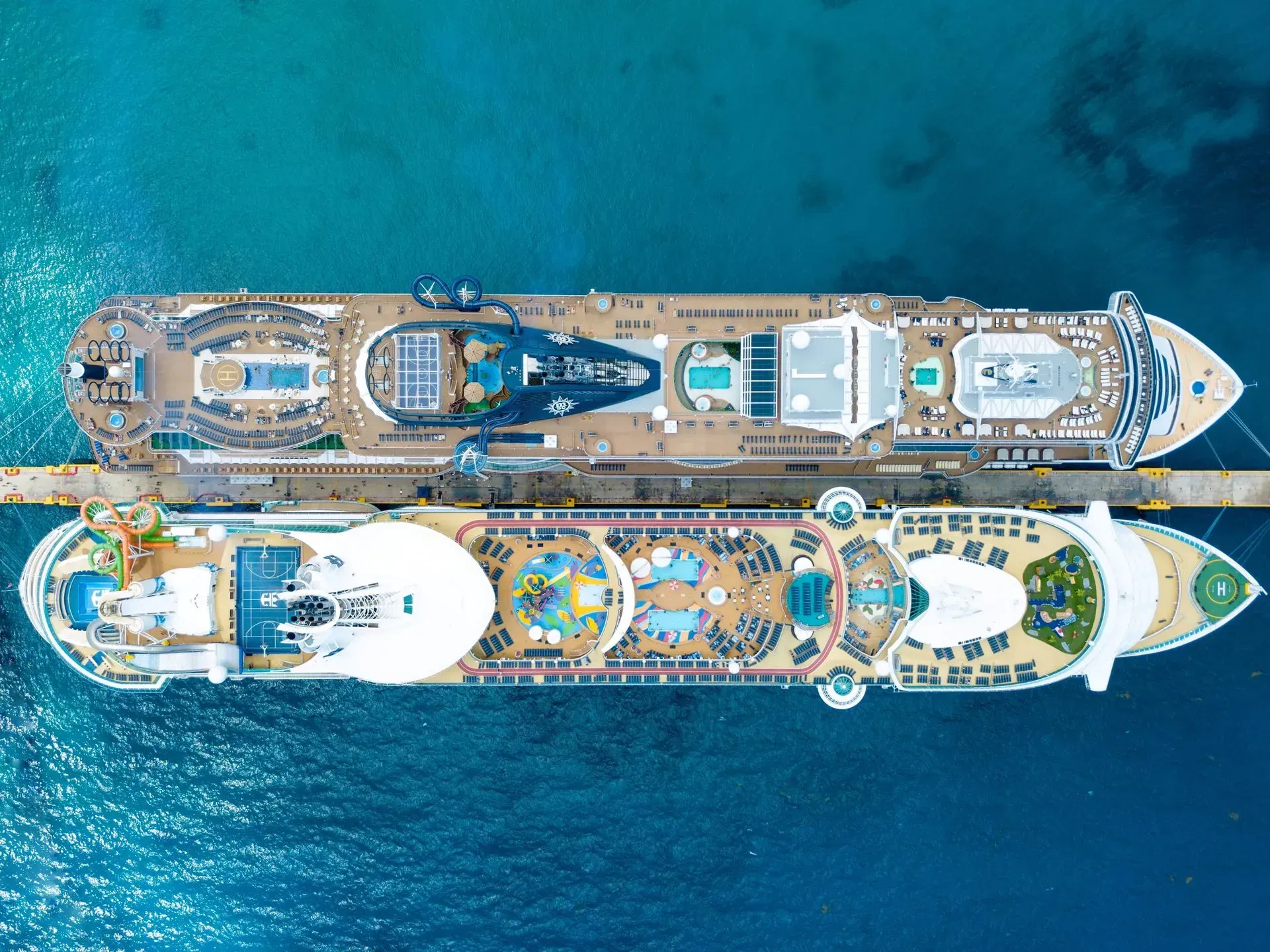 Two cruise ships docked side-by-side at a pier, seen from above. Bright blue water.