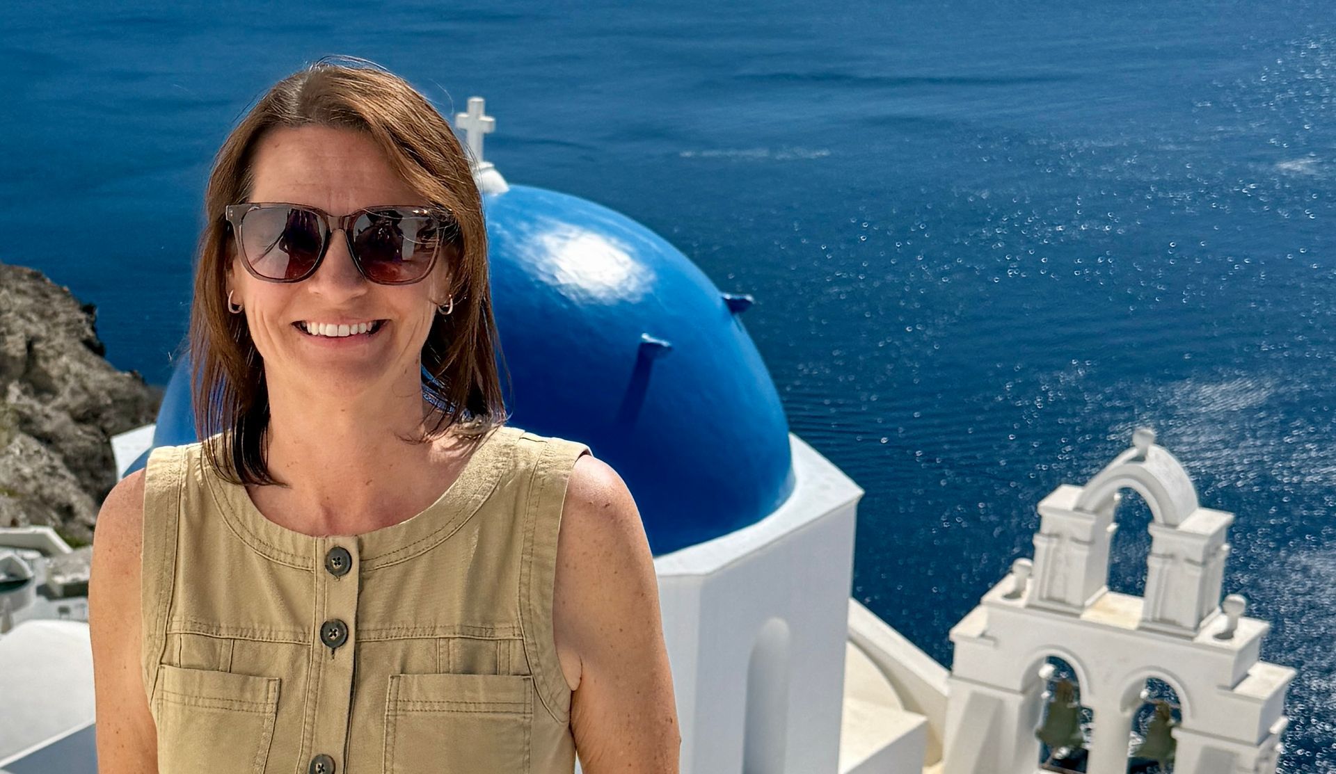 Woman wearing sunglasses, smiling, in front of white buildings with blue domes, overlooking the sea.