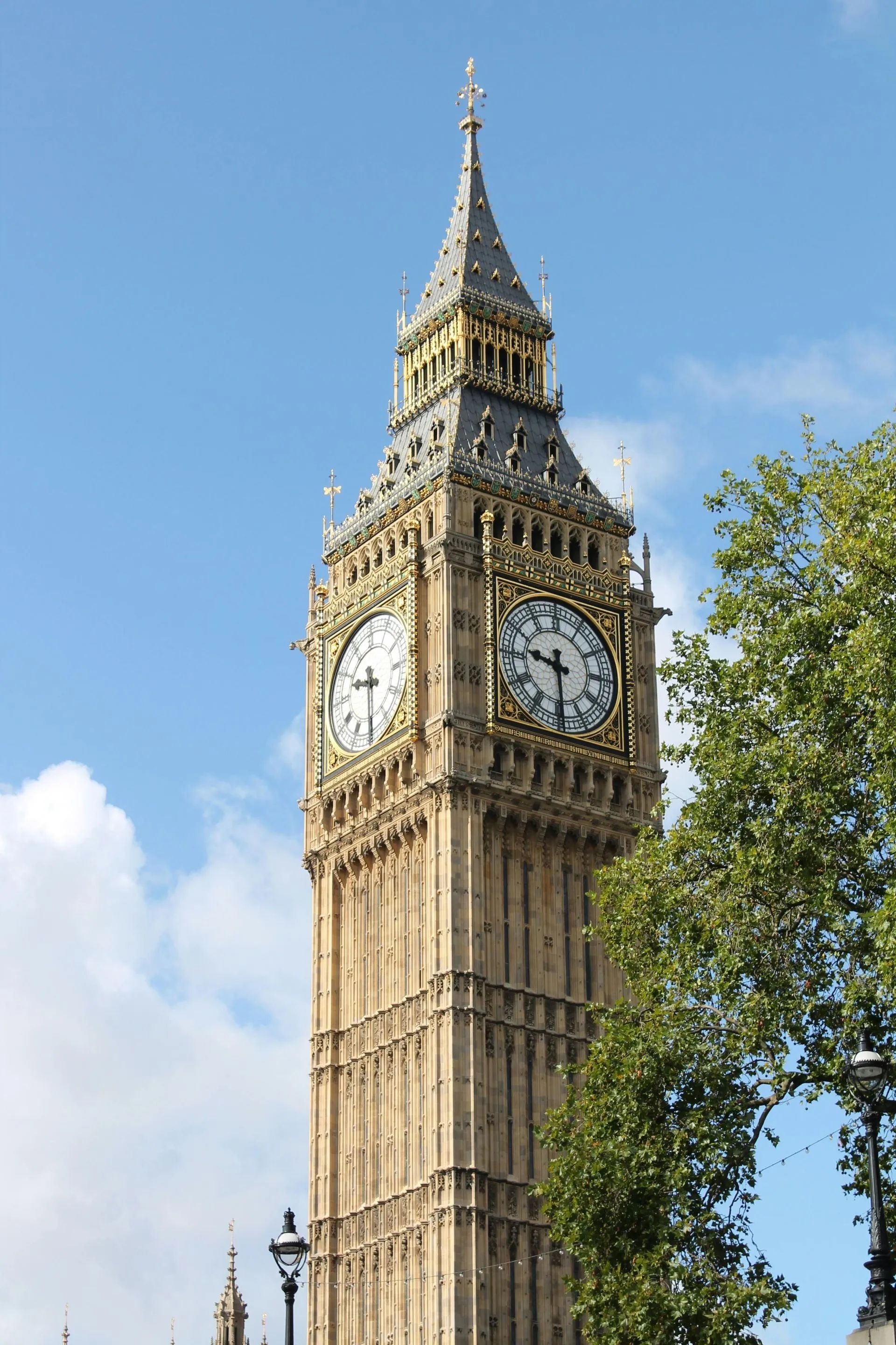 Clock tower, Big Ben, Westminster, London, with clock faces, against a blue sky, partially obscured by a tree.
