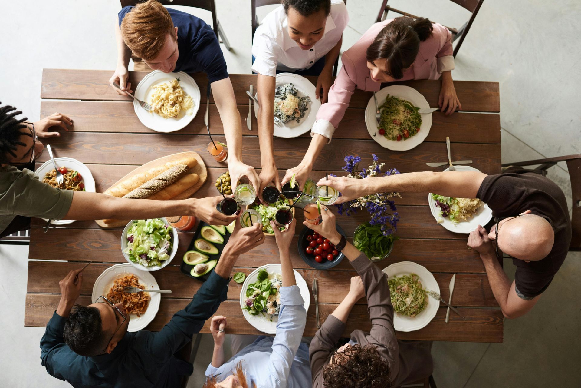 People at a wooden table toasting with glasses. Dishes of food, bread, and flowers are on the table.