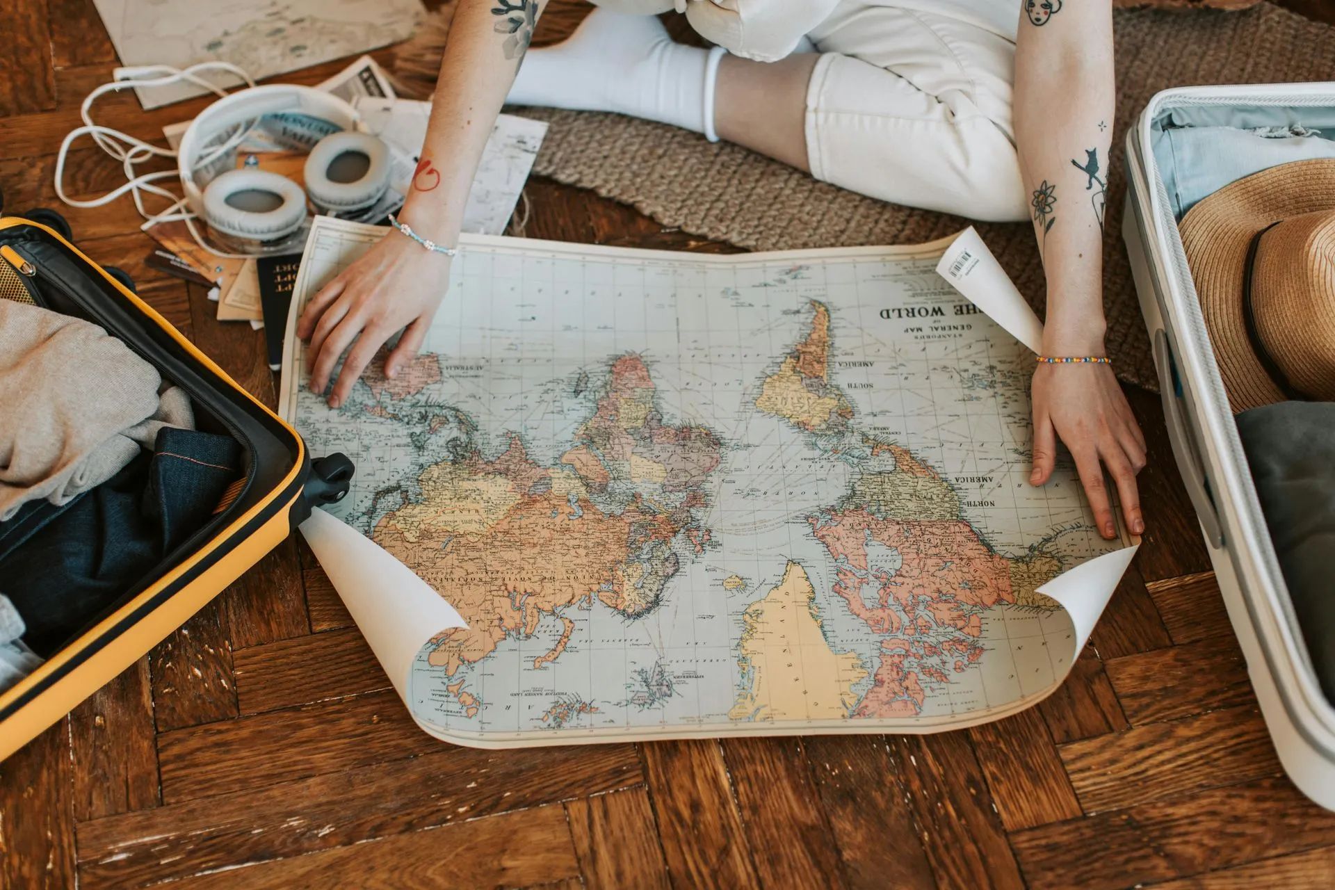 Person unfolds a world map on a wooden floor near open suitcases and packing items.
