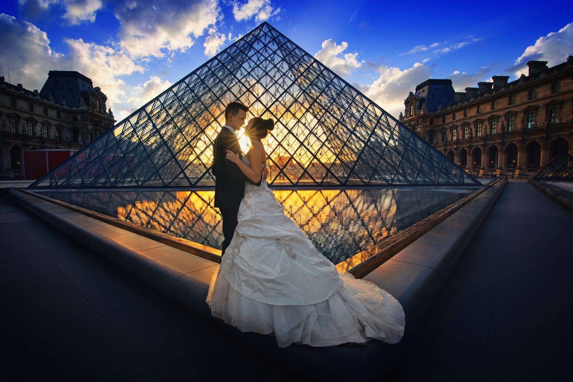 Couple in wedding attire embrace at the Louvre Museum in Paris, with the glass pyramid and setting sun.