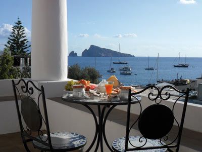 La colazione viene servita su un tavolo apparecchiato in una terrazza sul mare, con vista su una baia tranquilla e su un'isola in lontananza.