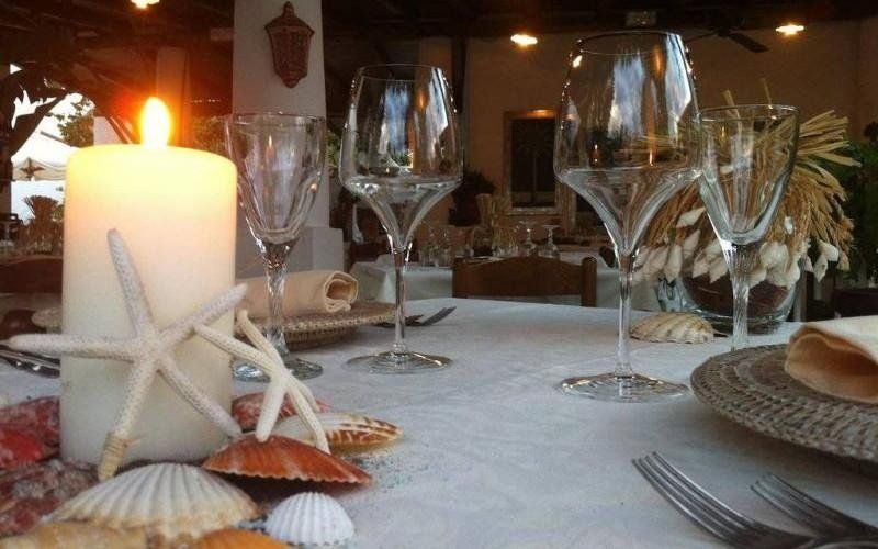 Tavola apparecchiata a lume di candela in riva al mare con stelle marine e conchiglie su una tovaglia bianca, bicchieri da vino e piatti.