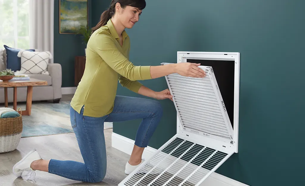 A woman is kneeling down in a living room to remove a filter from a vent.