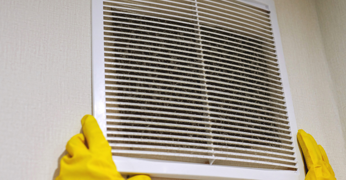 A person wearing yellow gloves is cleaning a vent on a wall.