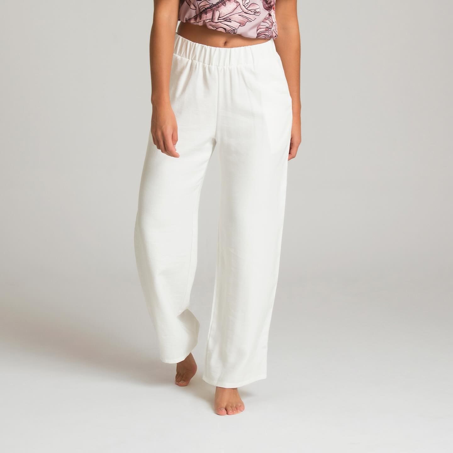 Mujer que viste pantalones blancos de pierna ancha y una blusa estampada, de pie descalza frente a un fondo blanco.