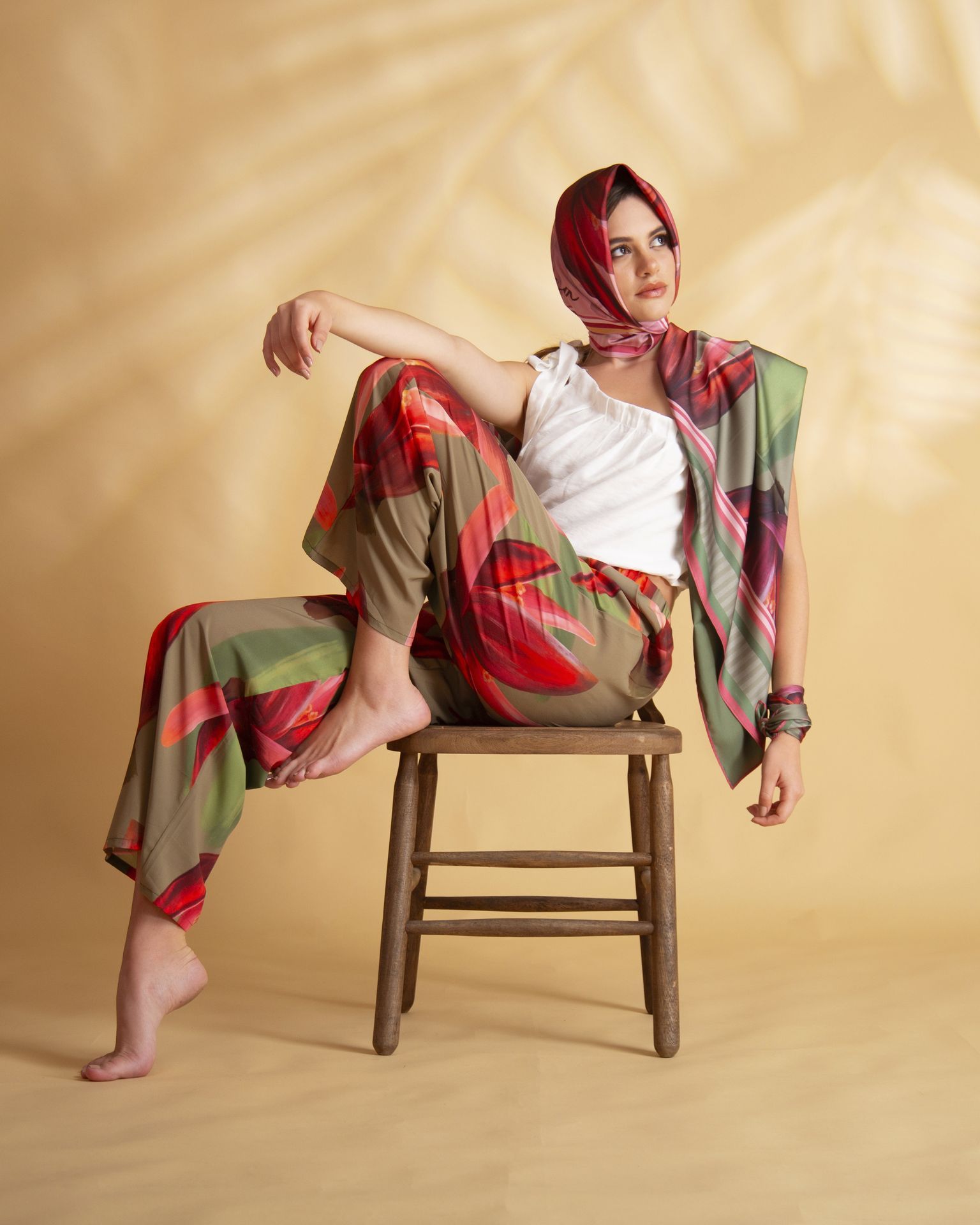 Una mujer con pantalones estampados y pañuelo en la cabeza está sentada descalza en un taburete de madera, mirando hacia otro lado. Lleva una blusa blanca y un chal.