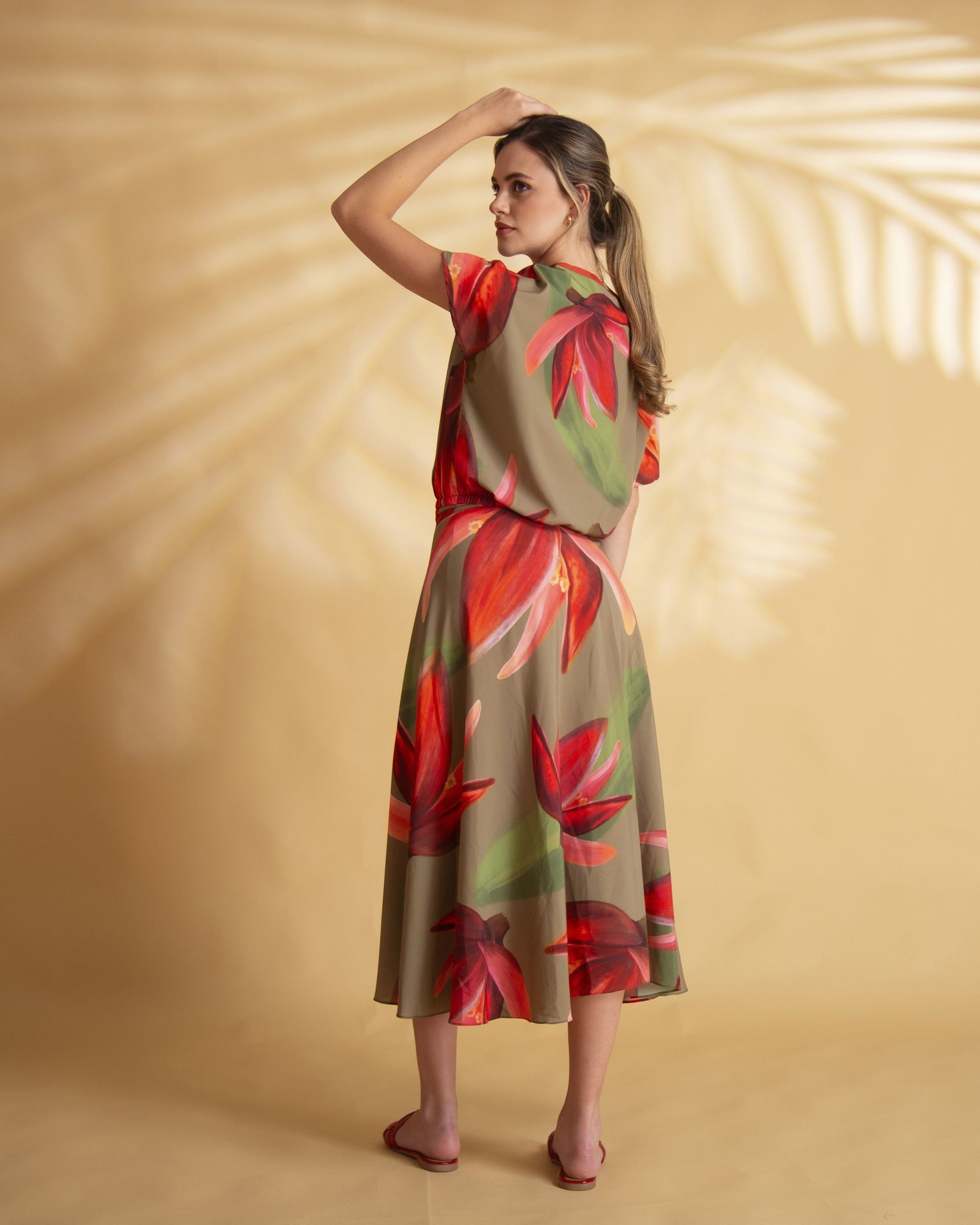 Una mujer con un vestido con estampado floral posa con el brazo levantado, de pie frente a un fondo beige con sombras de hojas.