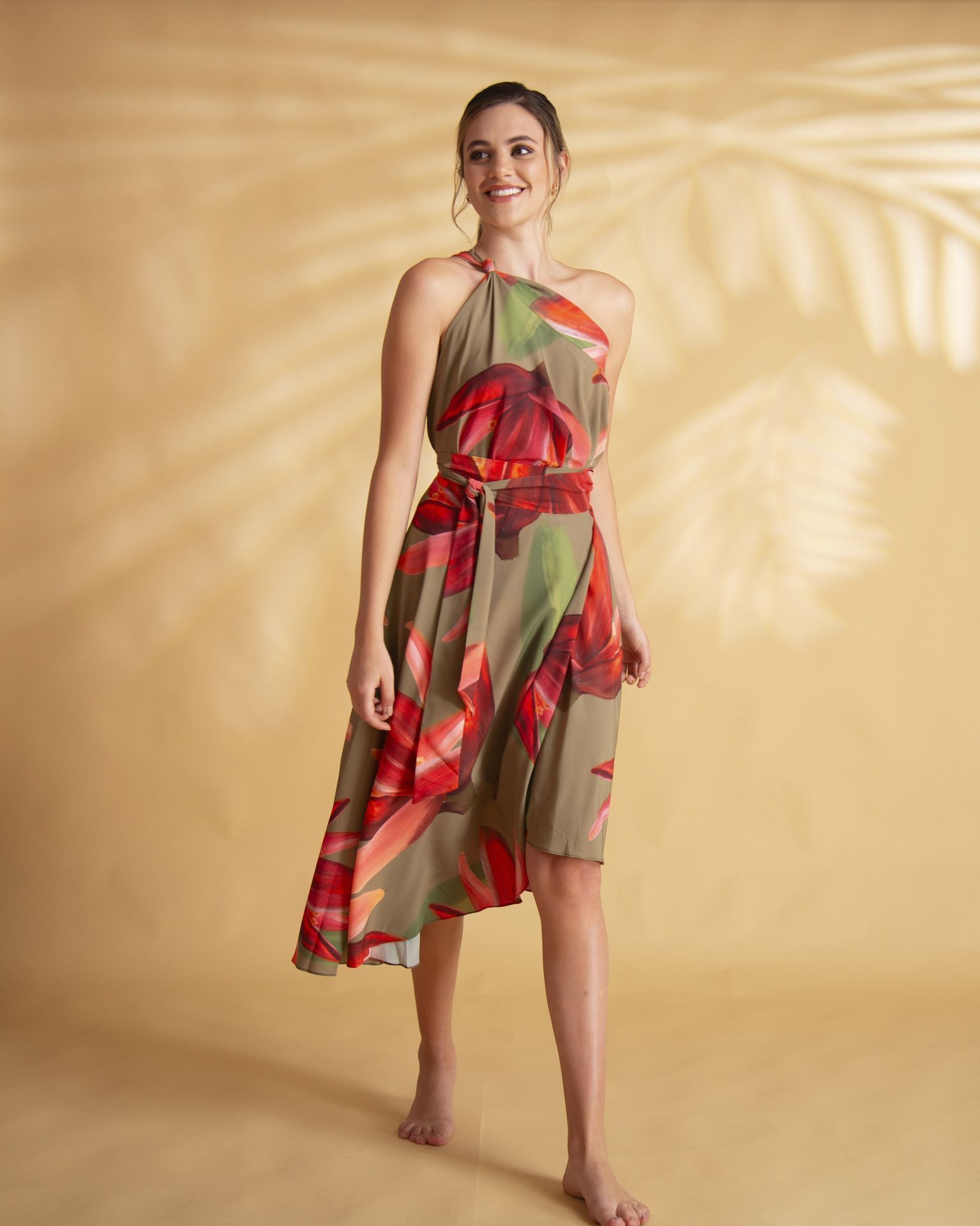 Mujer con un vestido halter floral, de pie frente a un fondo marrón claro con la sombra de una hoja. Sonríe.