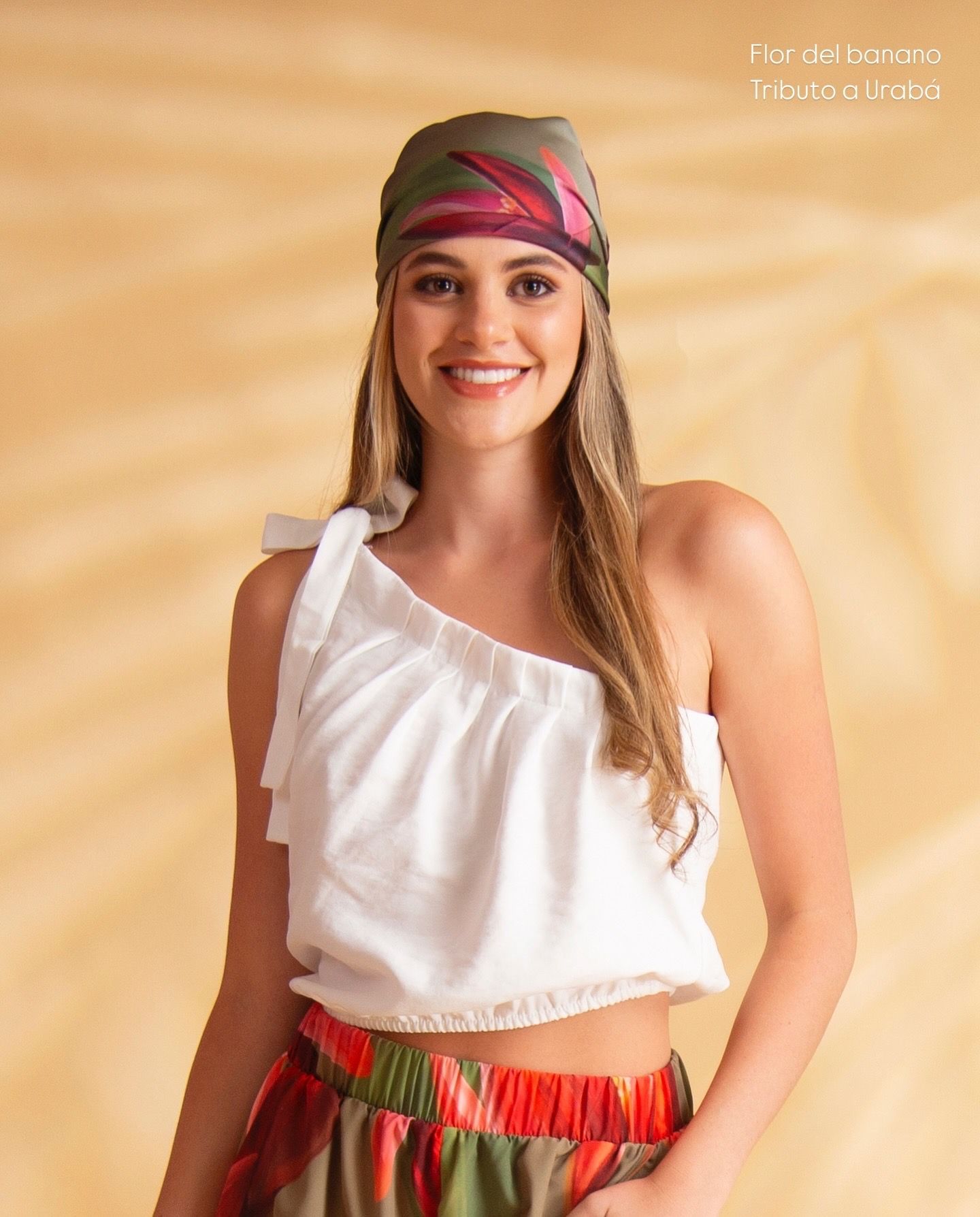 Mujer con blusa blanca de un solo hombro y pañuelo estampado, sonriendo. Posa sobre un fondo amarillo claro.