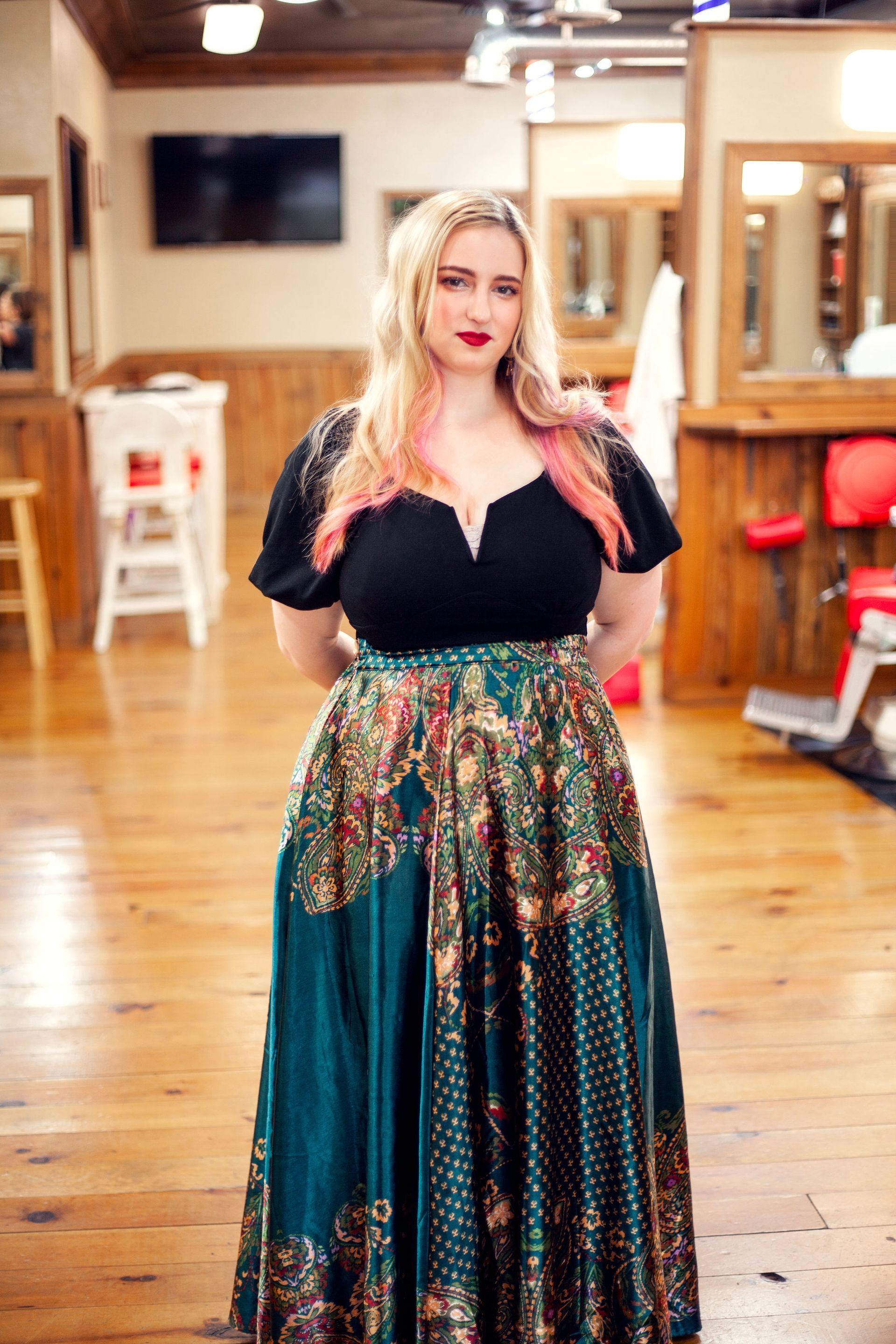 Woman with pink-streaked blonde hair, wearing a black top and patterned teal skirt, stands in a wood-paneled barber shop.