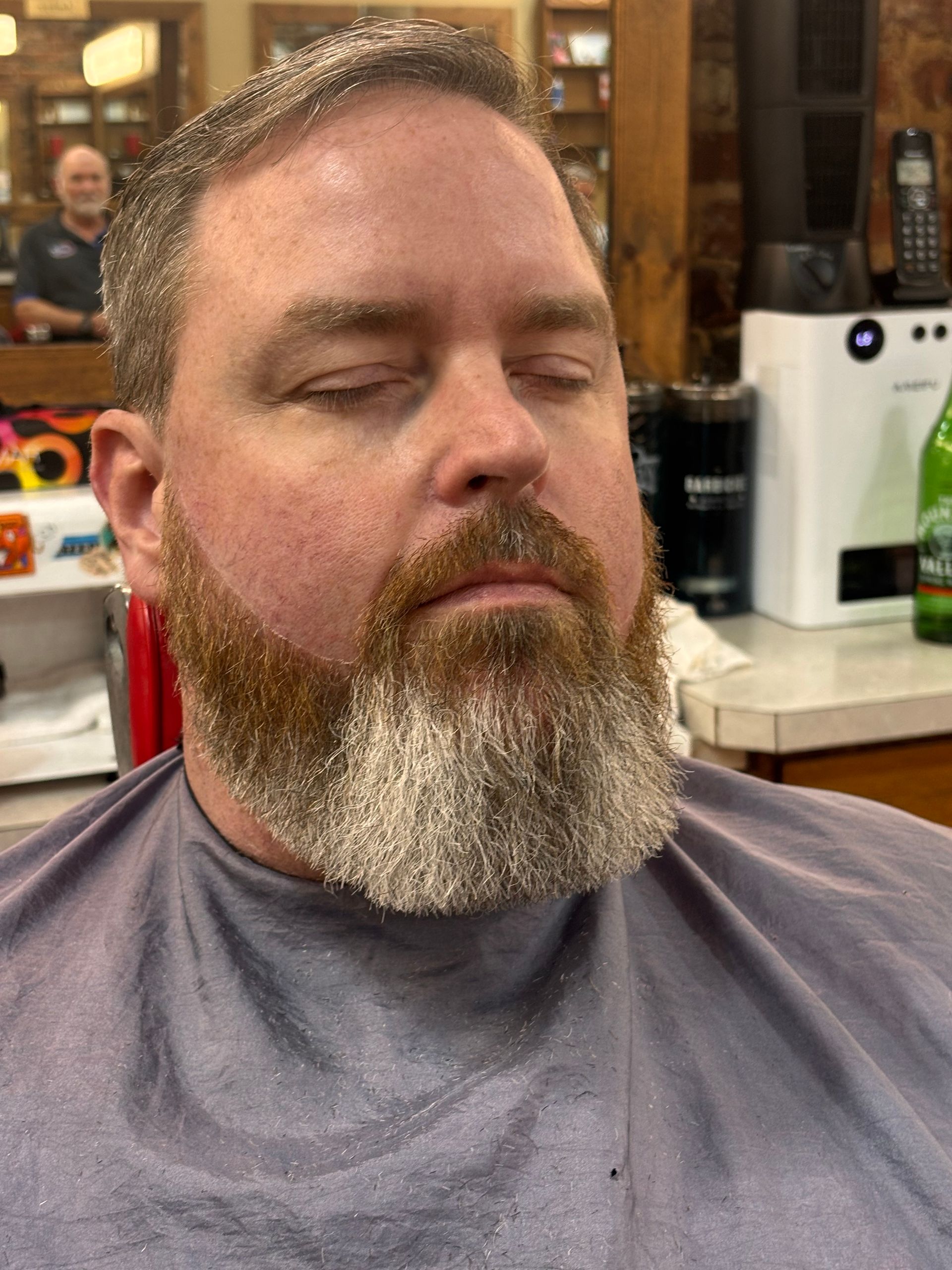 Man with closed eyes in a barber's chair, beard covered in snow-like material. Inside a barber shop.