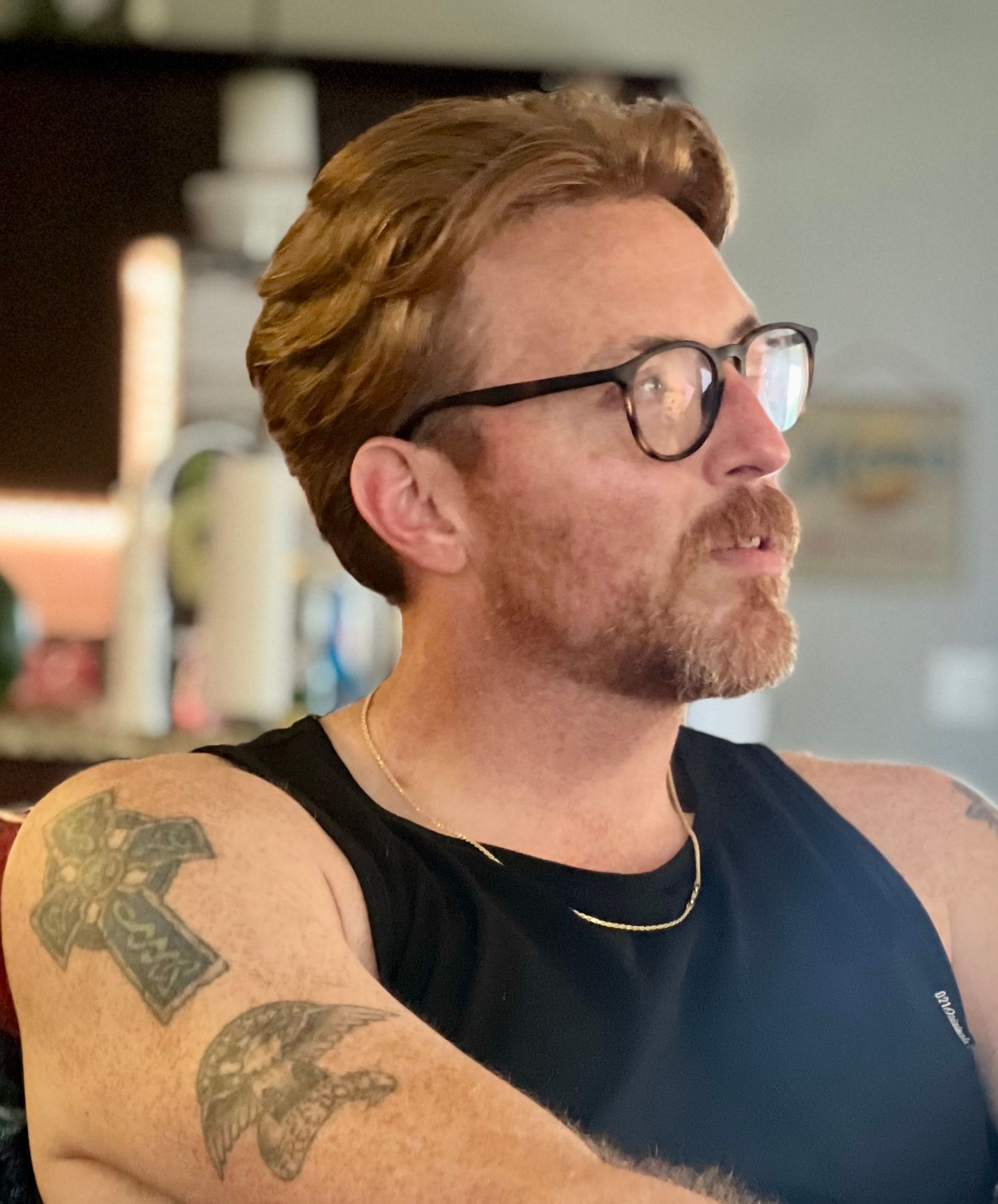 Man with reddish hair, glasses, and tattoos. Wearing a black tank top, looking right.