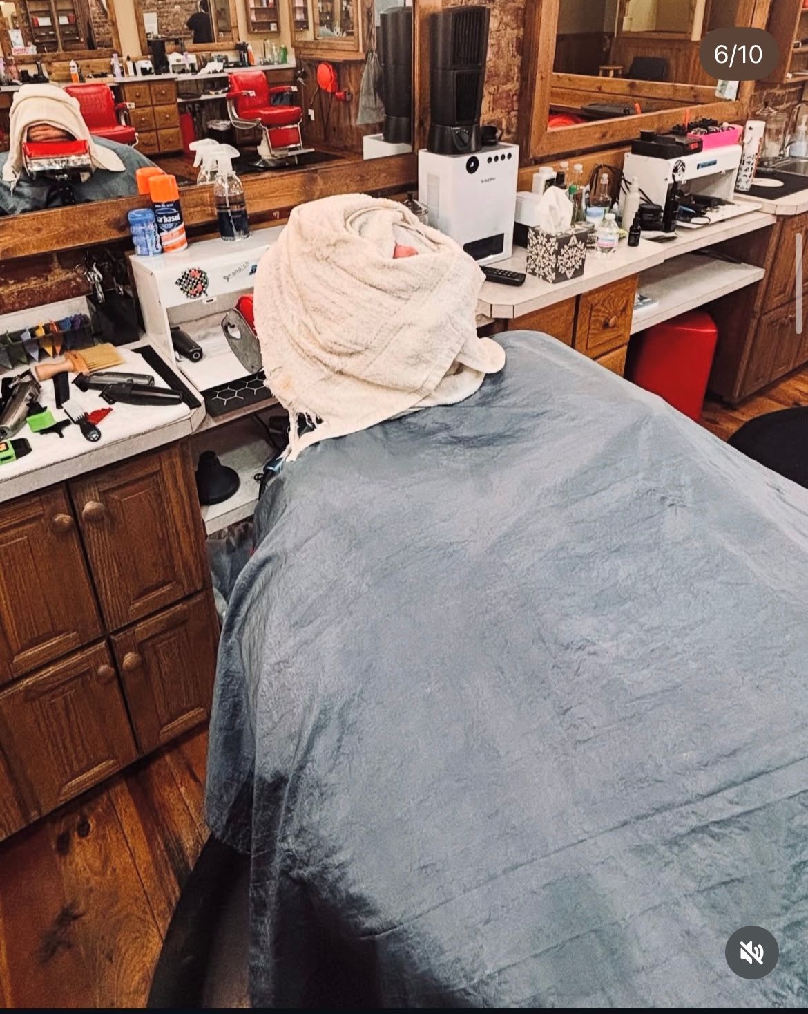 Person in barber chair with towel on head, covered by a blue cape, in a barbershop.