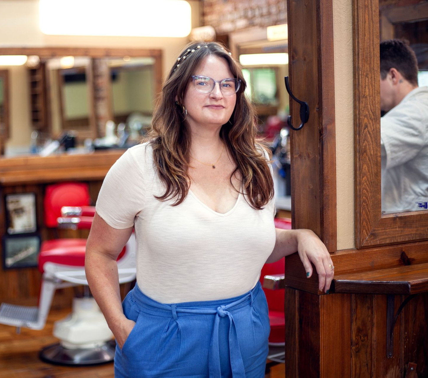 Woman in a barbershop, wearing glasses and blue pants, leaning on a wooden cabinet; a person is visible in the mirror.