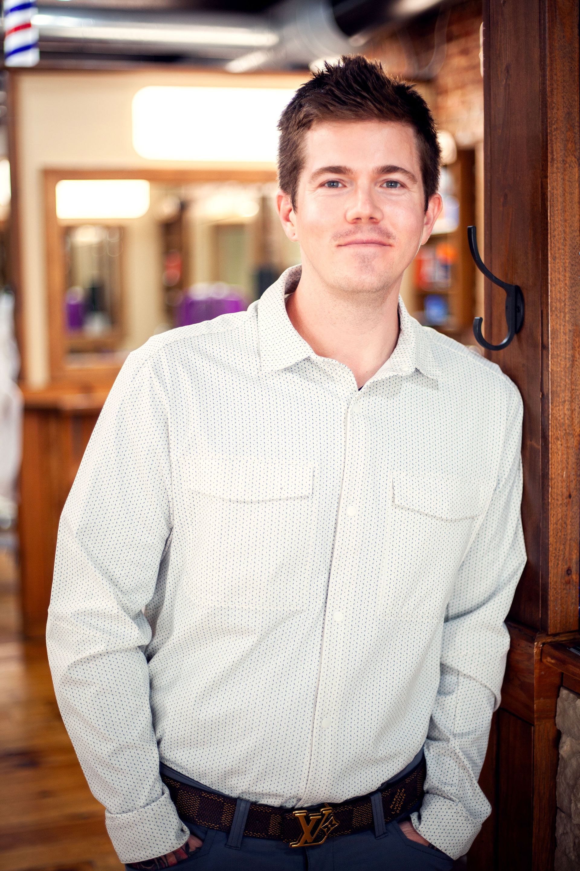 Man in a patterned shirt leans against a wooden post in a barber shop; looking at the camera.
