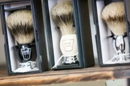 Three shaving brushes in black boxes, various handle styles, natural bristle color, on wooden shelf.