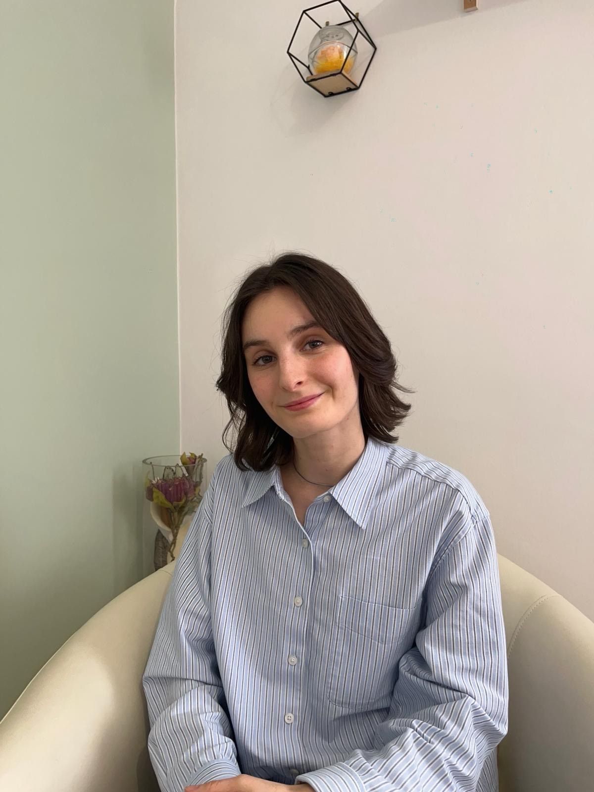 Una persona siede a braccia conserte in un ufficio, con un computer portatile sulla scrivania e un dipinto a pannelli multipli raffigurante un albero appeso alla parete.