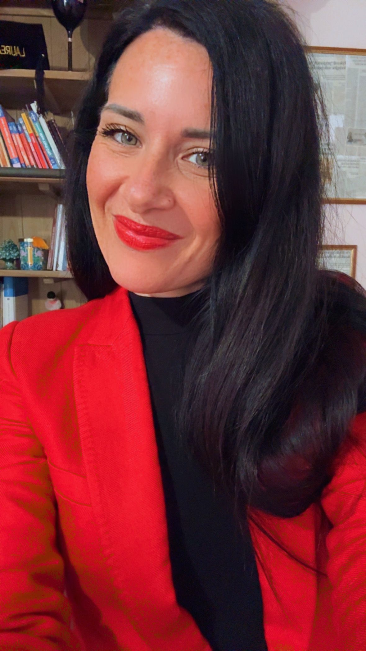 In una stanza con degli scaffali, una persona dai lunghi capelli scuri sorride indossando un blazer rosso acceso e un dolcevita nero.