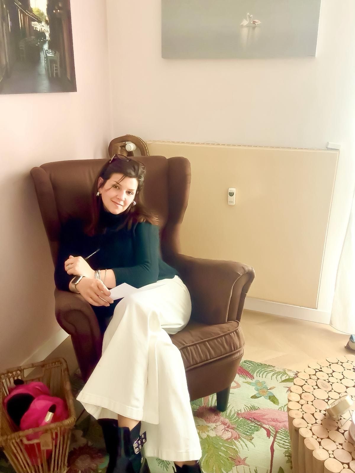 In una stanza ben illuminata, una persona con un dolcevita nero e pantaloni bianchi a gamba larga è seduta su una poltrona marrone con schienale alto.