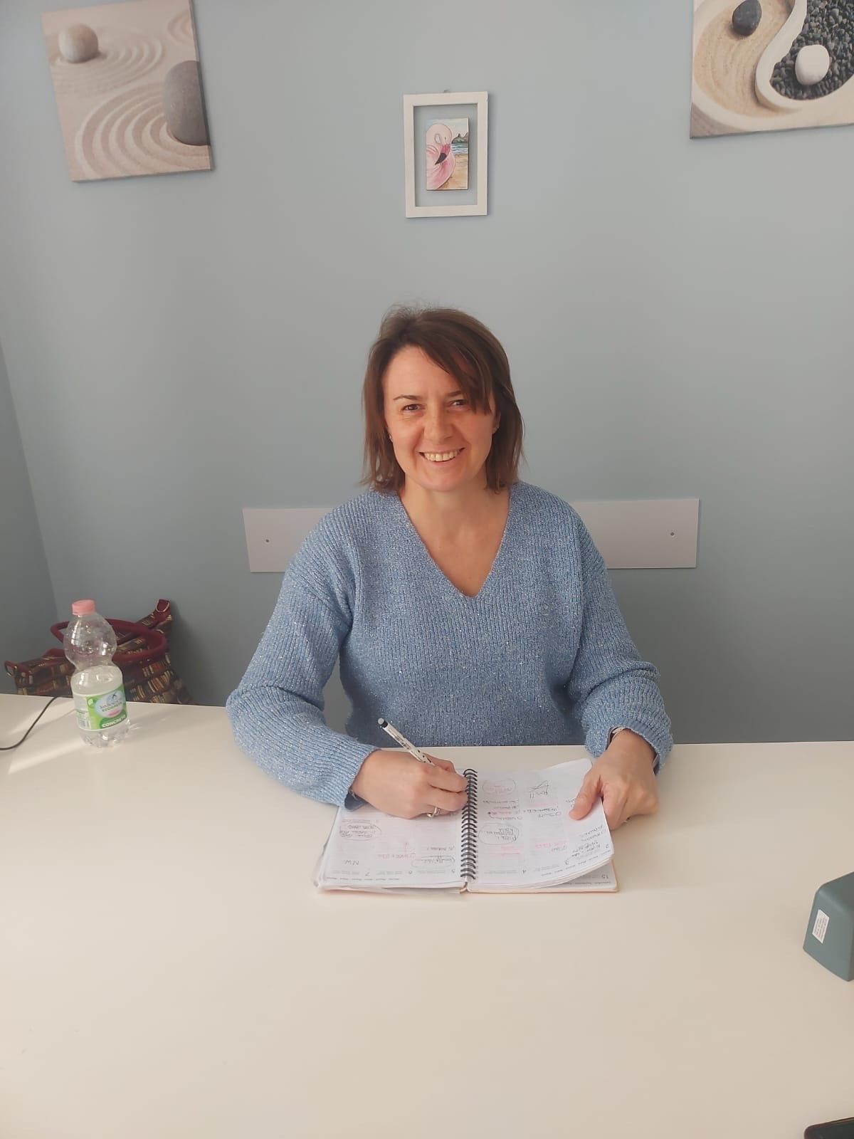 Una persona con lunghi capelli castani, che indossa un maglione a fantasia color crema con frange, è seduta a una scrivania appoggiata a una parete chiara.