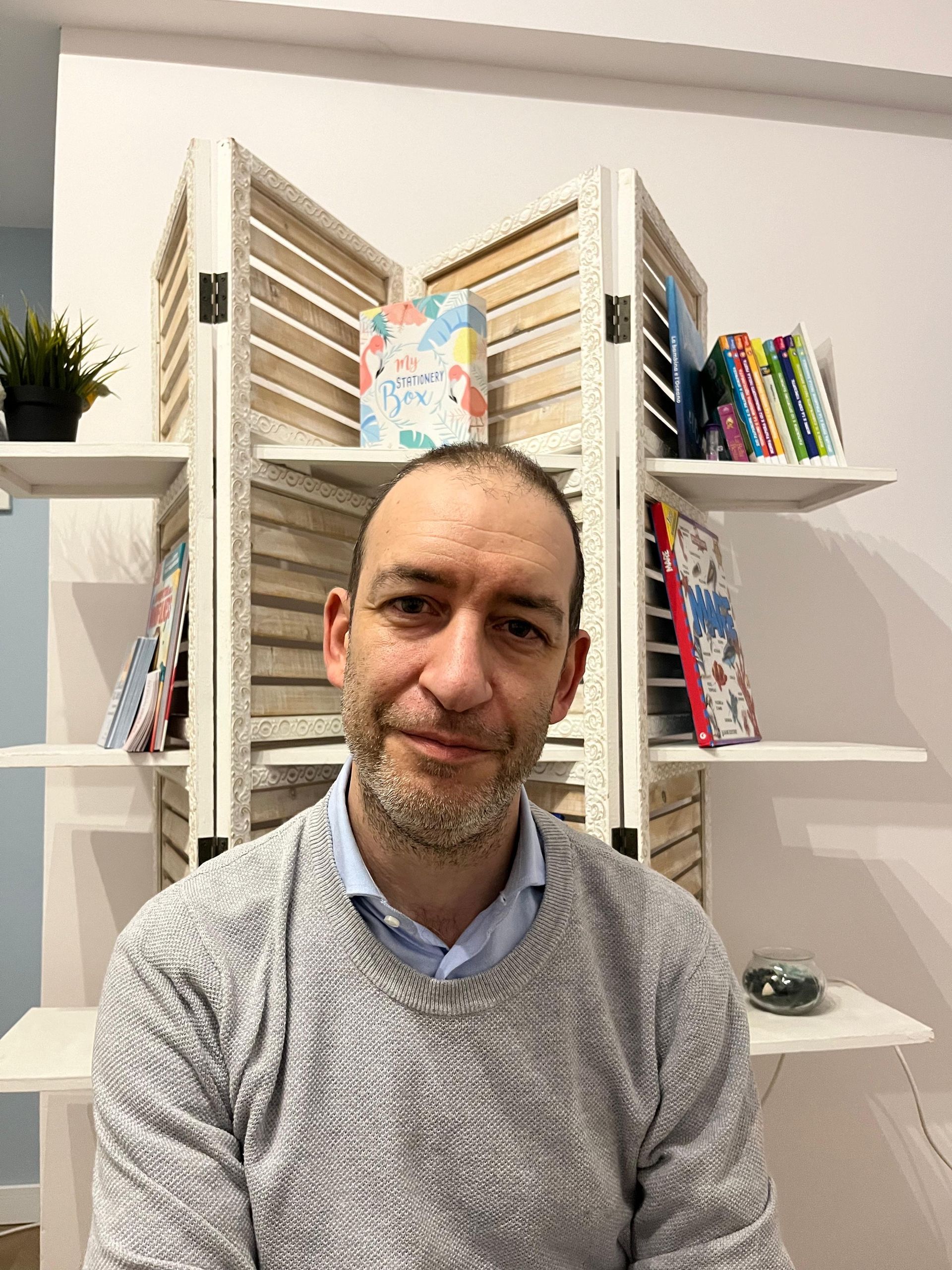 Una persona dai capelli scuri sorride mentre indossa una camicia a righe blu e bianche, seduta su una sedia.