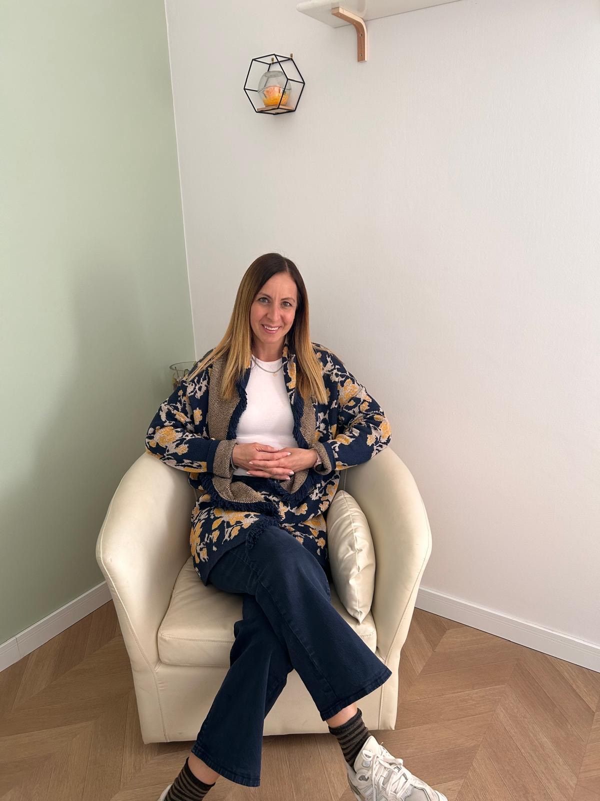 Una persona con i capelli biondi ondulati e gli occhiali sorride appoggiando il mento sulla mano, indossando un maglione nero.