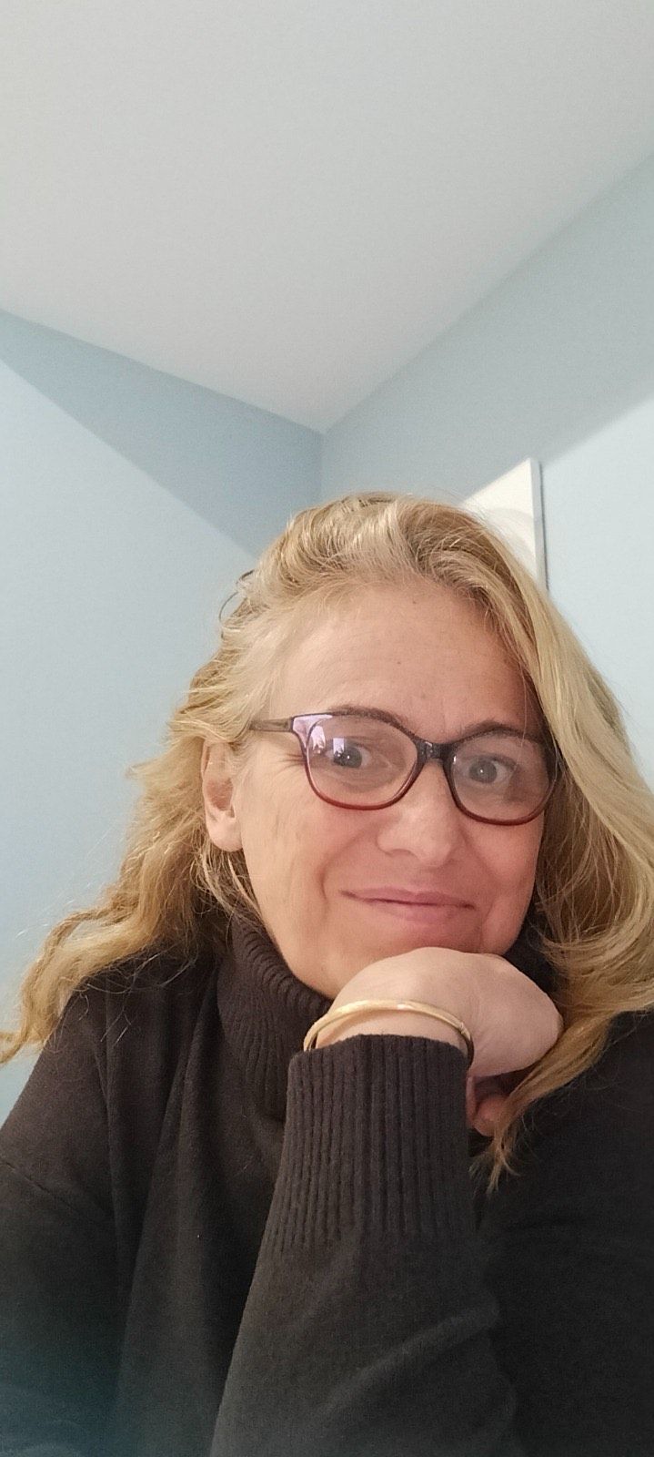 Una persona sorridente con indosso una giacca a fantasia è seduta su una poltrona color crema, appoggiata a una parete bianca e verde chiaro.