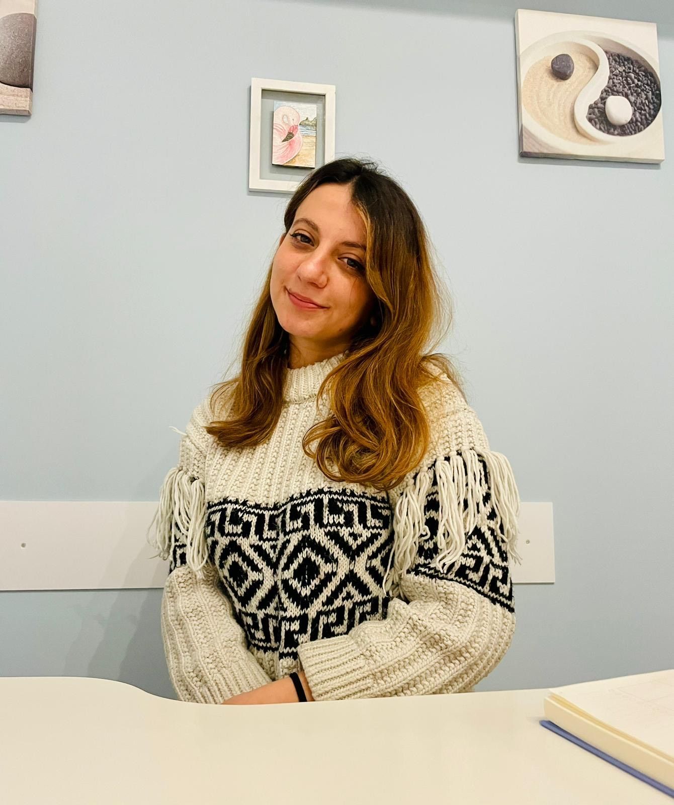 In una stanza azzurra, una persona con un maglione blu è seduta a una scrivania bianca e sorride mentre scrive su un quaderno a spirale.