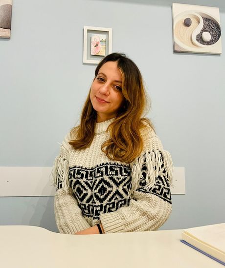 In una stanza azzurra, una persona con un maglione blu è seduta a una scrivania bianca e sorride mentre scrive su un quaderno a spirale.