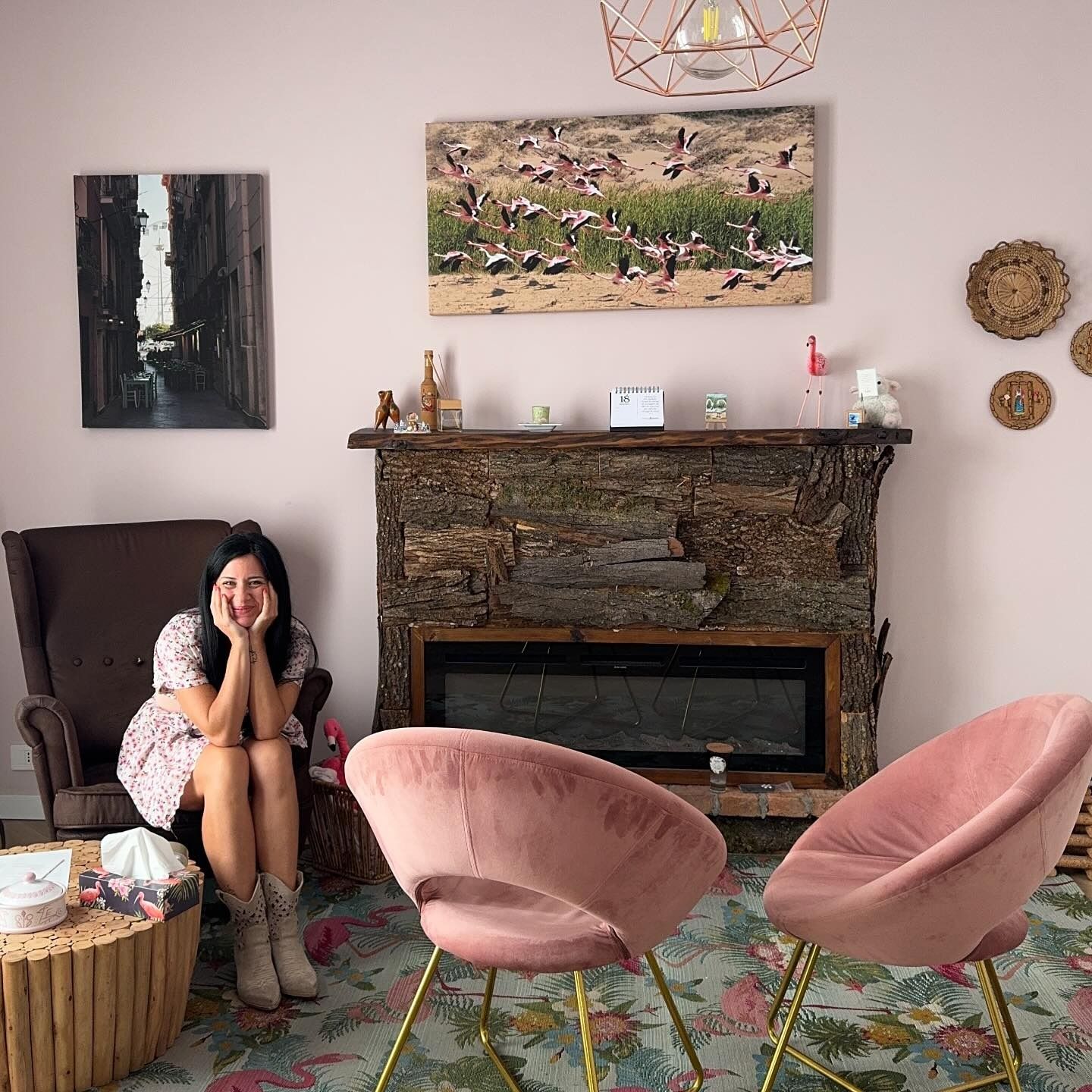 Una donna siede su una poltrona marrone accanto a un camino in pietra in una stanza con pareti rosa, due sedie rosa e un tappeto a fiori.