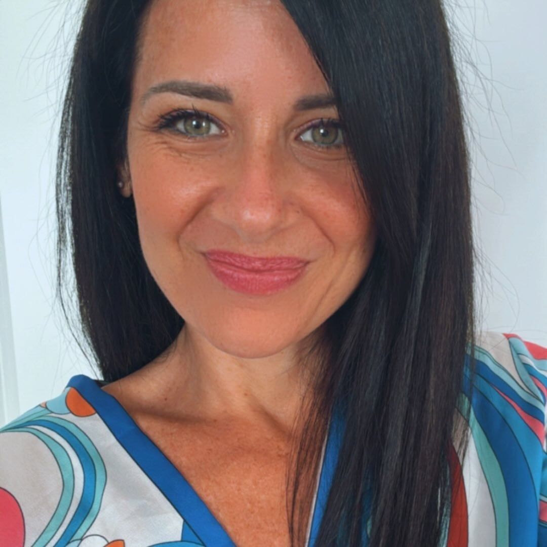 Una persona con lunghi capelli scuri sorride alla telecamera, indossando una maglietta a fantasia blu, bianca e rossa dai colori vivaci.