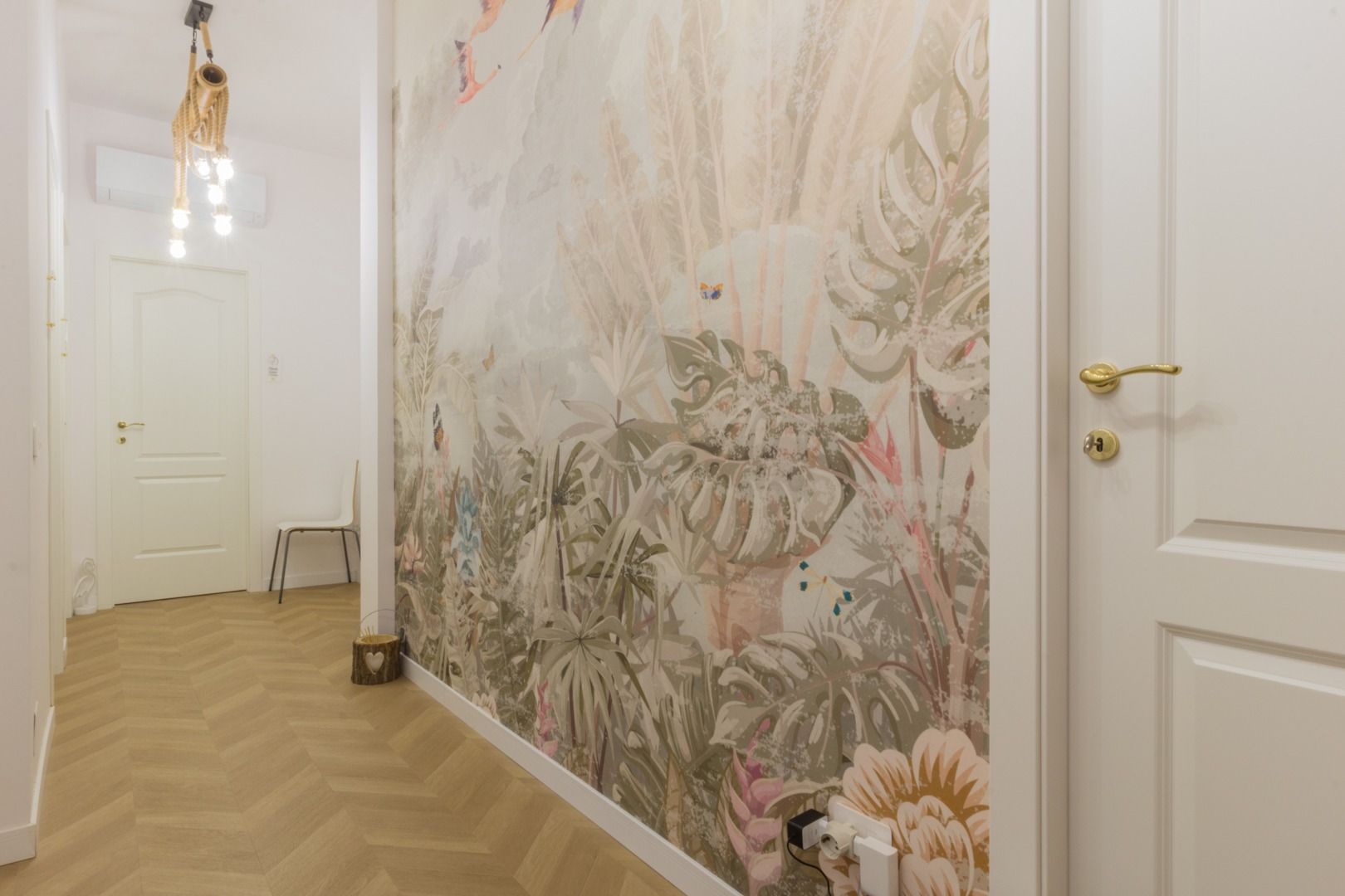 Un corridoio con pavimento a spina di pesce in legno chiaro, un murale raffigurante foglie tropicali, una lampada a sospensione e porte a pannelli bianchi.