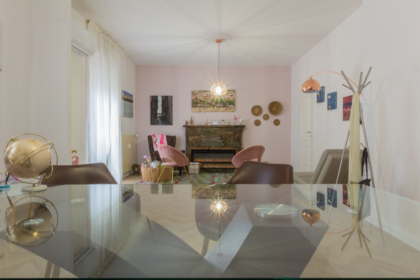 Un moderno soggiorno-sala da pranzo con un tavolo di vetro in primo piano, pareti rosa, poltrone e un camino decorativo.