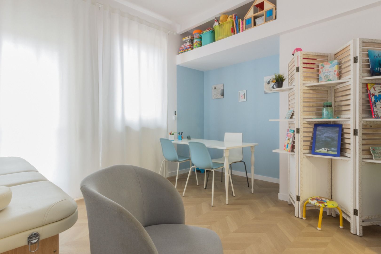 Una stanza dei giochi per bambini con un angolo azzurro, un tavolo e sedie, giocattoli su uno scaffale alto e un divisorio.